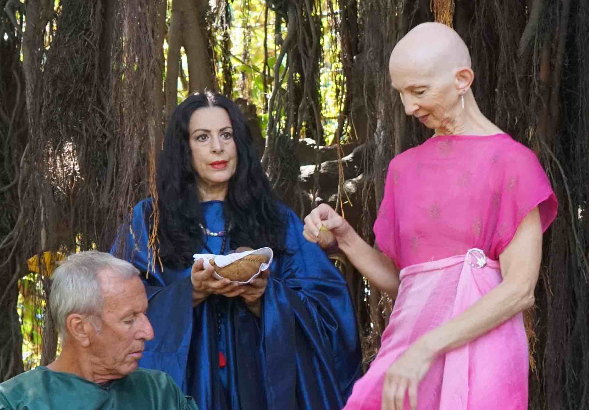 A striking woman in pink tempts a man in green with a fig under the gaze of a second woman wearing iridesant blue.