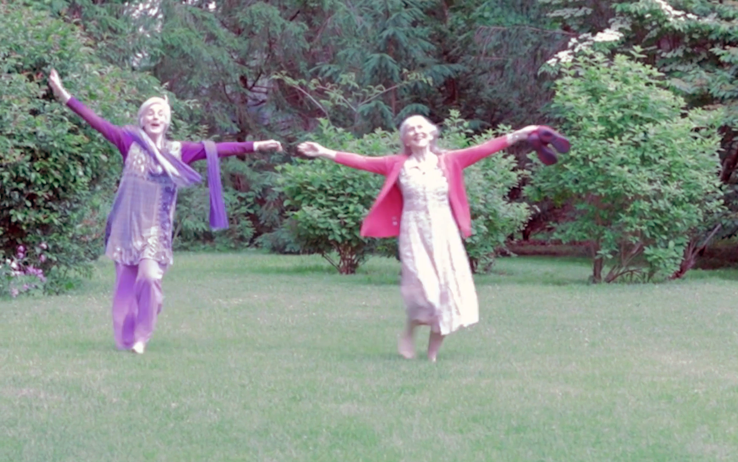 Two women dance in a garden with their arms outstretched.