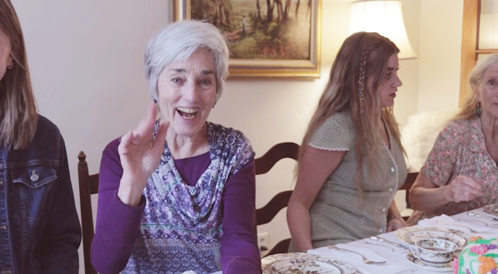 A woman raises her hand in greeting, sitting at a table set for afternoon tea