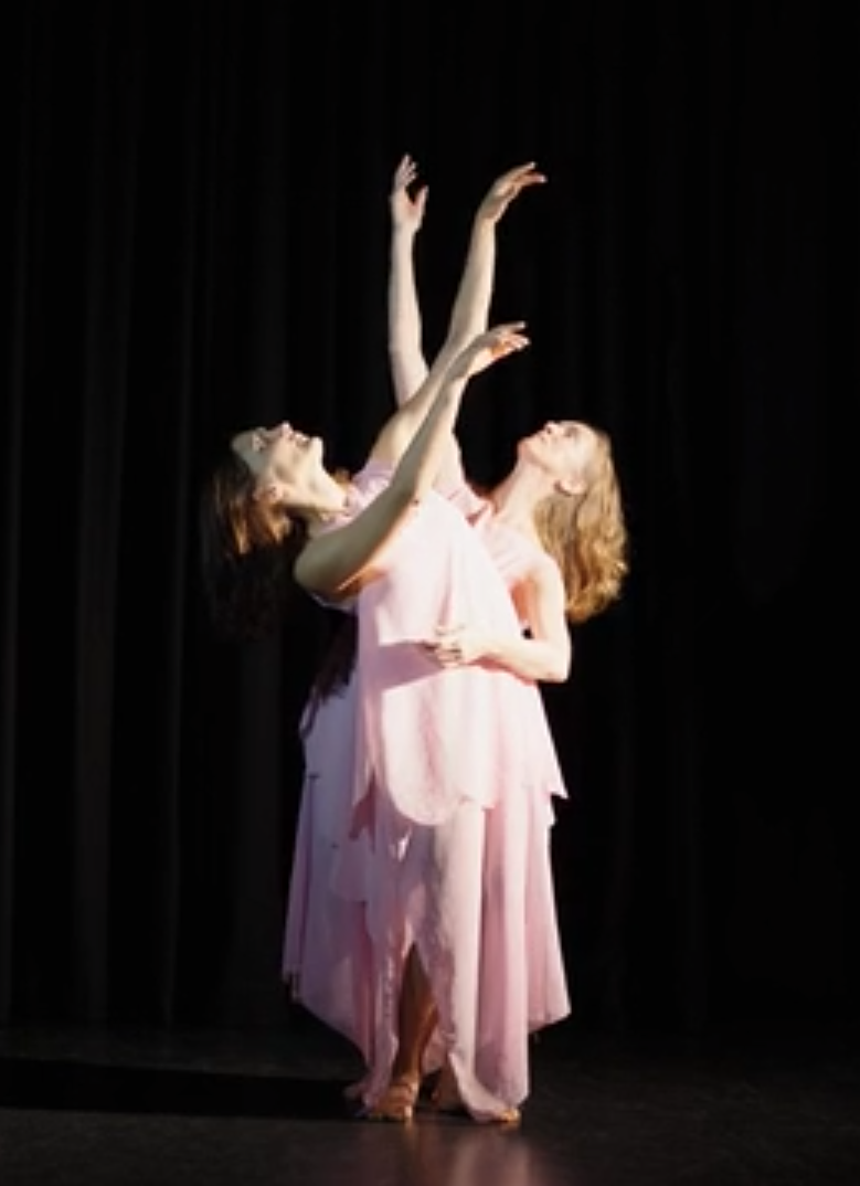 Two dancers in pink costumes against a dark background with arms raised