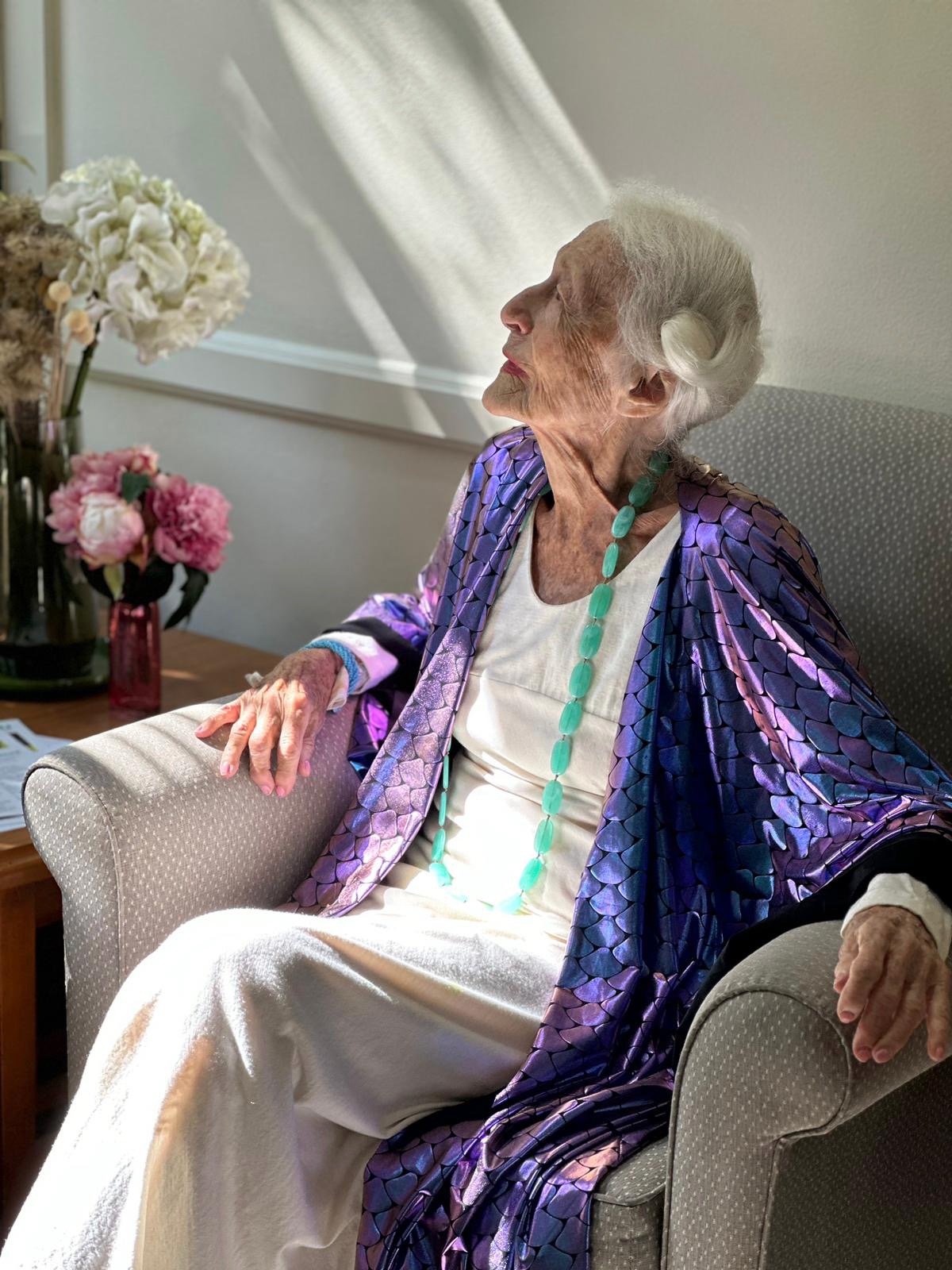 Eileen sitting in an armchair in the sun, wearing a purple coat.